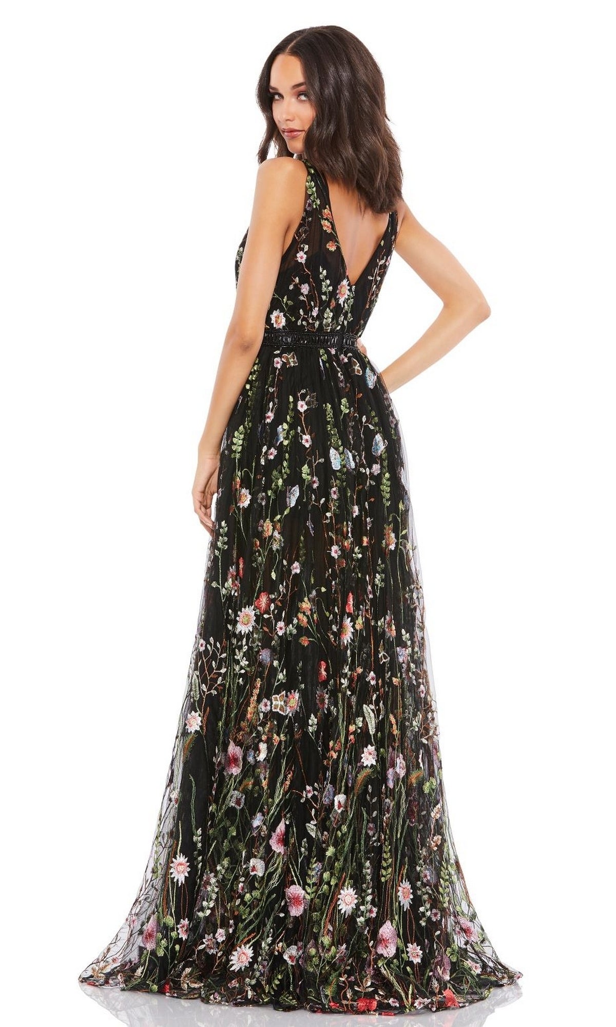 A woman faces away, wearing the Long Formal Dress 70132 by Mac Duggal—a sleeveless black floral gown with a deep V-back and colorful embroidery—standing against a plain white background.